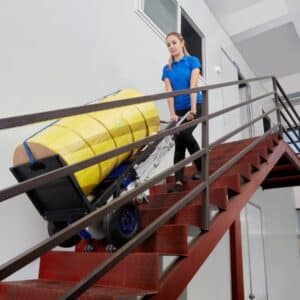 Stair climber rental with wheels transporting a strapped cylindrical load down industrial stairs, female operator controlling descent
