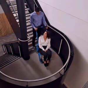 Man assisting a woman on a mobility chair on a curved staircase, showing stairlifts for narrow curved stairs in use.