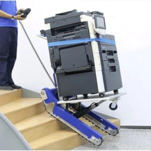 Electric stair trolley carrying a large office photocopier up wooden stairs, operated by a worker using handheld controls.