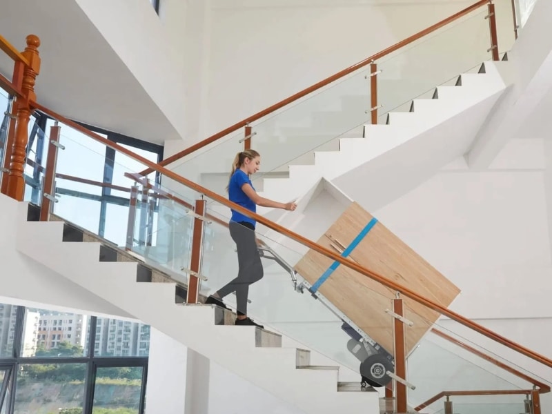 Heavy duty hand trolley carrying large wooden cabinet upstairs, operated by a woman using stair-climbing wheels indoors.